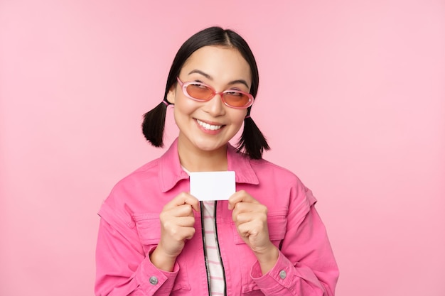 세련된 현대 아시아 소녀의 초상화는 할인 신용 카드를 보여주고 분홍색 배경 위에 서 있는 비접촉식 쇼핑 개념을 지불하는 것을 기쁘게 생각합니다.
