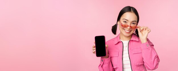 Portrait of stylish modern asian girl shows mobile phone screen smartphone app interface standing in sunglasses against pink background