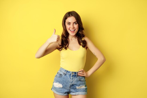 Portrait of stylish modern girl, feminine woman showing thumbs up, recommending gesture, like or approve, praise, standing over yellow background