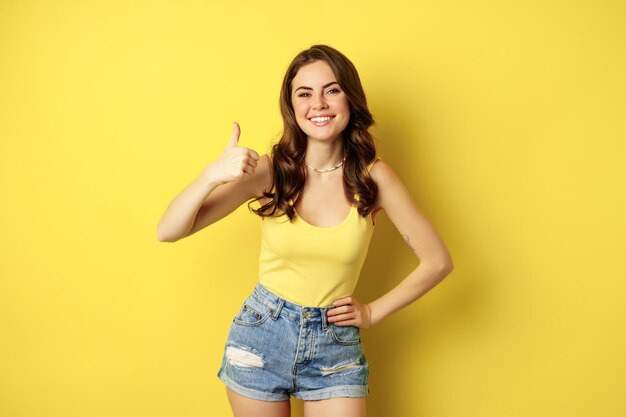 Portrait of stylish modern girl, feminine woman showing thumbs up, recommending gesture, like or approve, praise, standing over yellow background