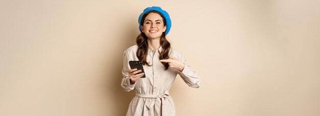 Portrait of stylish modern woman in outerwear pointing at mobile phone screen and looking happy smil