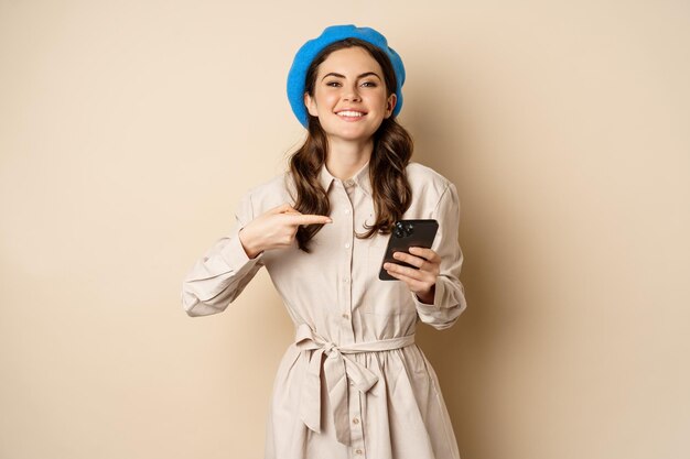 Portrait of stylish modern woman in outerwear pointing at mobile phone screen and looking happy smil...