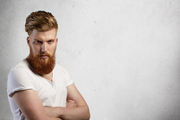 Portrait of stylish redhead hipster with fuzzy beard wearing white t-shirt with rolled up sleeves posing indoors with arms crossed, having sullen expression on his face. 