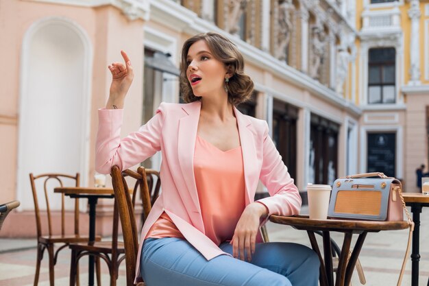 Portrait of stylish sensual lady sitting at table drinking coffee in pink jacket summer style trend, blue handbag, accessories, street style, women fashion