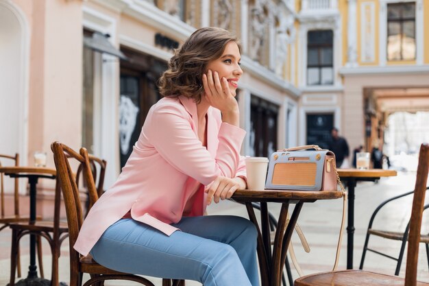 Портрет стильной улыбающейся дамы, сидящей за столом, пьющей кофе в розовой куртке, летний стиль, тренд, синяя сумочка, аксессуары, уличный стиль, женская мода