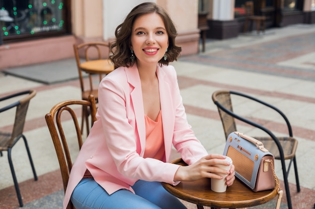 Портрет стильной улыбающейся дамы, сидящей за столом, пьющей кофе в розовой куртке, летний стиль, тренд, синяя сумочка, аксессуары, уличный стиль, женская мода