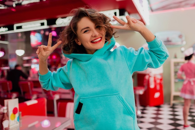 Portrait of stylish smiling woman in blue hoodie in cafe having fun in cheerful mood trendy fashion style