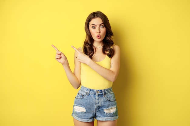 Portrait of stylish summer girl, brunette woman showing way, pointing fingers left, advertising, standing over yellow background