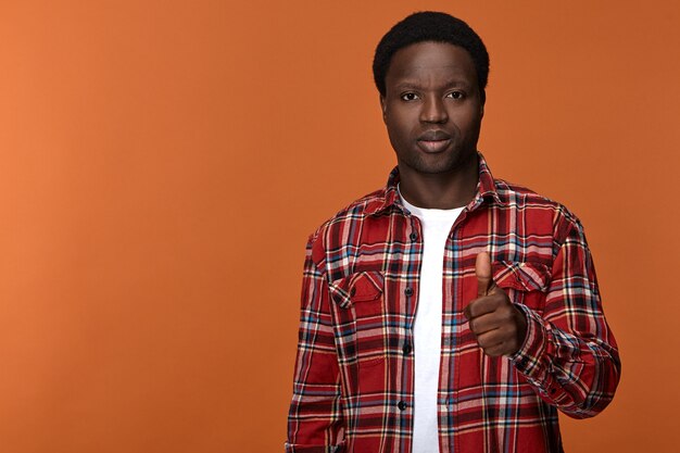 Portrait of stylish trendy looking young Afro American man in his twenties posing against blank  wall with thumbs up gesture, expressing approval, satisfaction and positive attitude