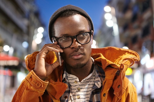 Free photo portrait of stylish young afro american male having nice conversation on mobile, spending evening outdoors with city lights