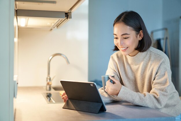 Portrait of stylish young asian woman watching videos on tablet sitting in kitchen and drinking coff