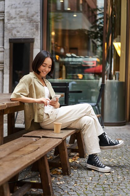 カフェに座っているスタイリッシュな若い韓国人女性の肖像画は、スマートフォンの笑顔を保持し、屋外でコーヒーを楽しんでいます