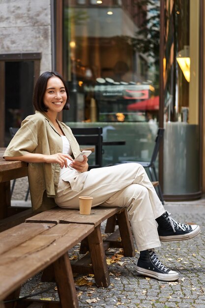 カフェに座っているスタイリッシュな若い韓国人女性の肖像画は、スマートフォンの笑顔を保持し、屋外でコーヒーを楽しんでいます