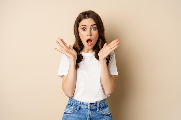 Free photo portrait of stylish young woman gasping, looking surprised and amazed, impressed by smth, standing in t-shirt and jeans over beige background