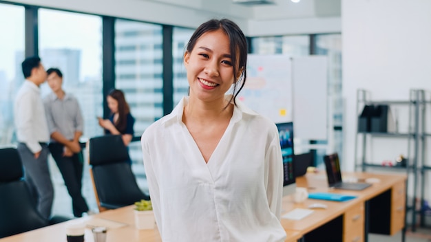 Portrait of successful beautiful executive businesswoman smart casual wear looking at camera and smiling in modern office workplace. Young Asia lady standing in contemporary meeting room.