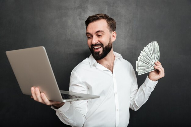 Portrait of successful businessman in white shirt holding fan of money dollar banknotes and silver notebook in both hands over dark gray