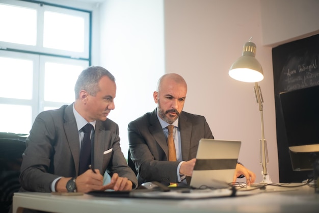 Free photo portrait of successful businessmen in office. two serious man in suits working with computer at table, making decisions and developing strategy for business growth. business growth and finance concept