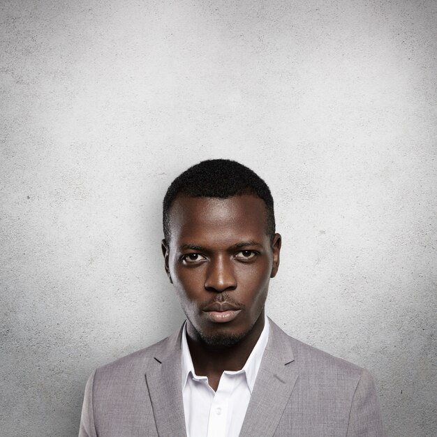 Portrait of successful confident dark-skinned entrepreneur wearing gray formal suit, standing against wall with  