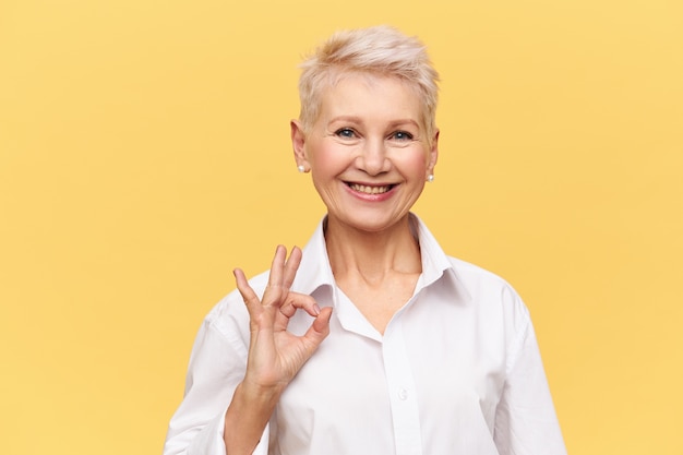 Portrait of successful confident middle aged businesswoman with short dyed hair with broad smile making ok gesture, rejoicing at good profitable deal and great yearly income
