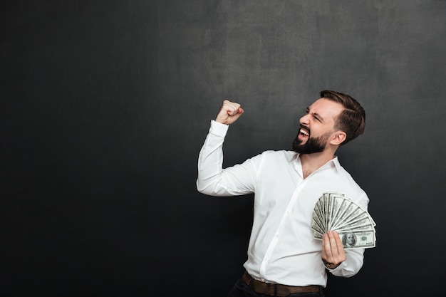 Portrait of successful guy in white shirt rejoicing like winner with fan of 100 dollar bills in hand, clenching fist aside over dark gray