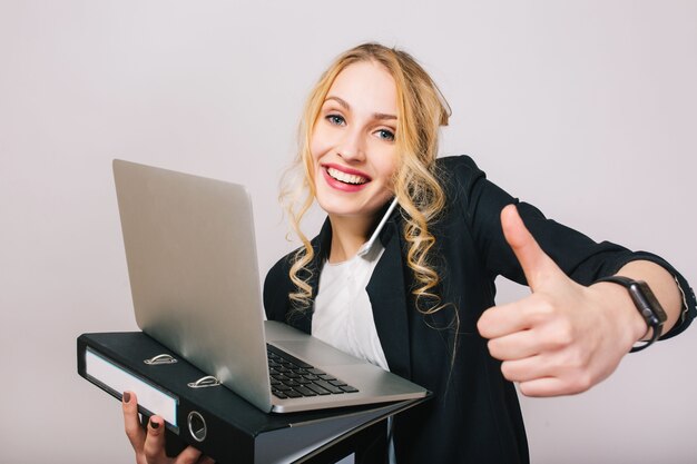 Portrait successful joyful businesswoman smiling, holding laptop, folder, talking on phone isolated. Wearing white shirt and black jacket, modern office worker