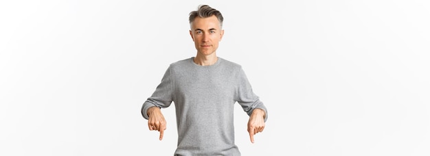 Free photo portrait of successful middleaged man in grey sweater pointing fingers down and looking confident at camera showing logo standing over white background