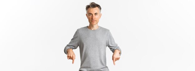 Portrait of successful middleaged man in grey sweater pointing fingers down and looking confident at camera showing logo standing over white background