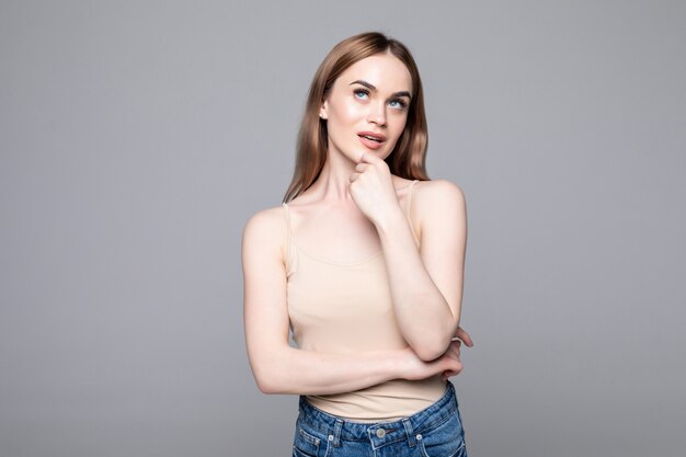 Portrait of successful woman looking aside and holding finger at cheek while smiling isolated over gray wall