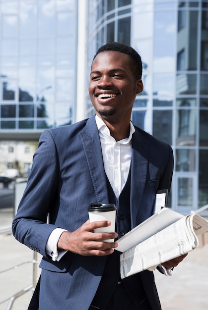 使い捨てのコーヒーカップを保持している成功したアフリカの若手実業家の肖像画。新聞とデジタルタブレット