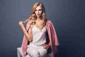 Free photo portrait of successful young seductive blonde woman in pink pastel jacket sitting on table