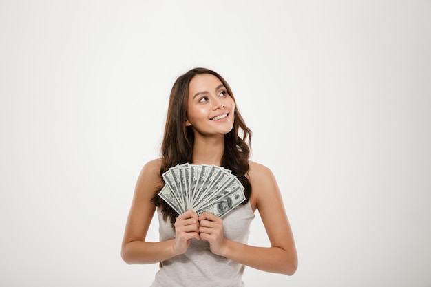 Portrait of successful young woman with long hair holding lots of money cash, smiling on camera over white wall