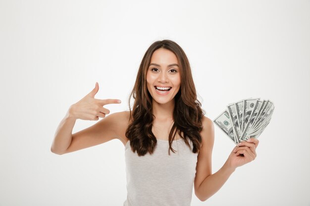 Portrait of successful young woman with long hair showing lots of money cash, smiling on camera over white wall