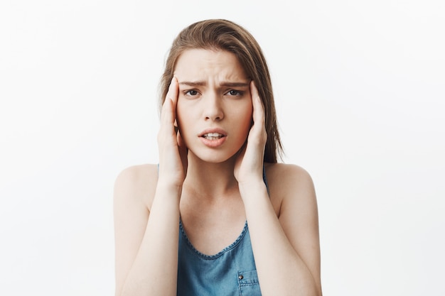 Portrait of suffering young attractive caucasian student girl with dark long hair in blue shirt massaging forehead with fingers,  with tired expression after long day at work