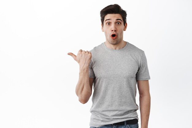 Portrait of surprised 30s man pointing left aside showing banner with amazed face say wow at camera standing over white background