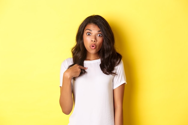 Portrait of surprised africanamerican girl pointing finger at herself standing over yellow background