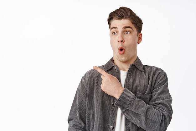 Portrait of surprised and amazed guy pointing, gasping and looking left on white