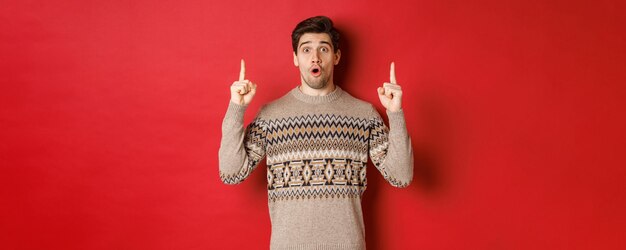 クリスマスのプロモーションのオファーを示し、クリスマスセーターを着て、赤い背景の上に立っている驚いて驚いたハンサムな男の肖像画。