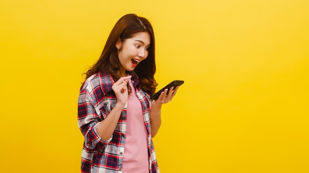 Portrait of surprised asian female using mobile phone with positive expression, dressed in casual clothing and looking at camera over yellow wall. happy adorable glad woman rejoices success.