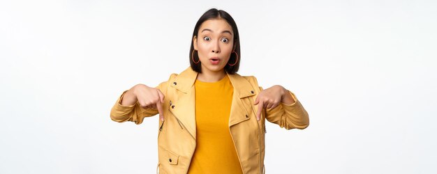 Portrait of surprised asian girl looking amazed and impressed pointing fingers down showing banner with shocked face expression white background