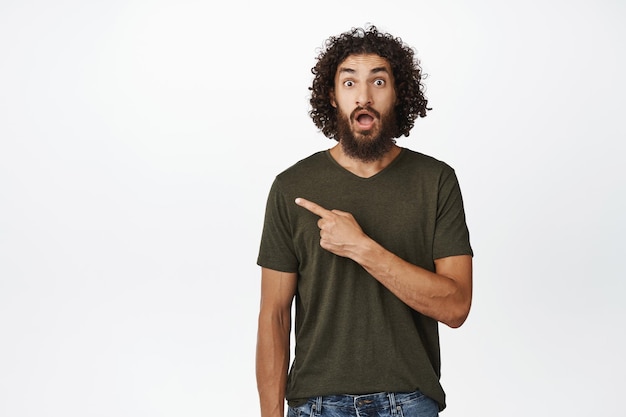 Portrait of surprised bearded middleeaster man pointing left drop jaw and look amazed at camera recommending link standing over white background