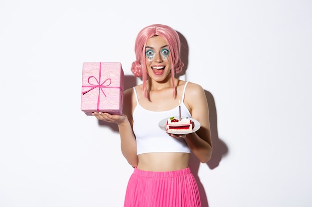Portrait of surprised beautiful girl in pink wig, receive birthday gift, holding b-day cake and smiling happy, standing.