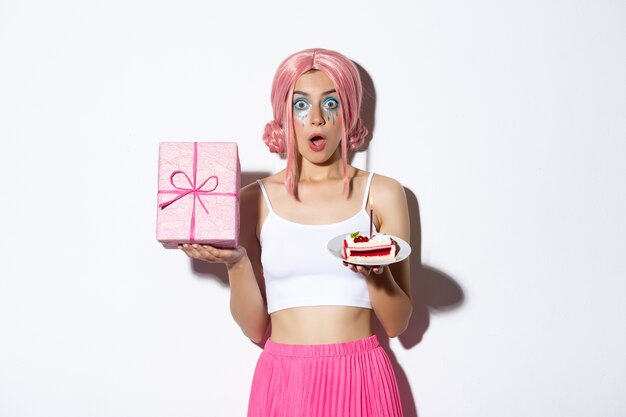 Portrait of surprised beautiful girl in pink wig, receive birthday gift, holding b-day cake and smiling happy, standing.