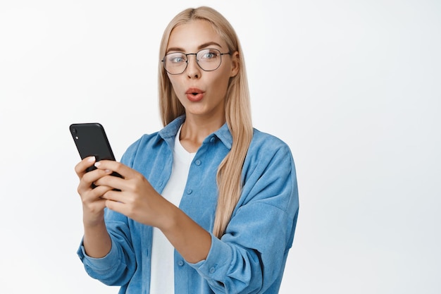 Портрет удивленной блондинки в очках, держащей смартфон и заинтригованно смотрящей в камеру, стоящую на белом фоне
