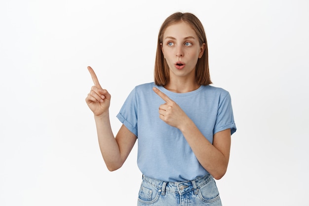 Portrait of surprised blond woman gasp, say wow, pointing and looking left at logo, showing sale advertisement or discount banner, standing over white wall.