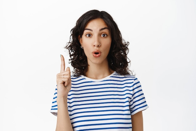 Portrait of surprised brunette modern girl, gasp and say wow, pointing up at advertisement, showing interesting promo deal, tell about discounts, standing  on white