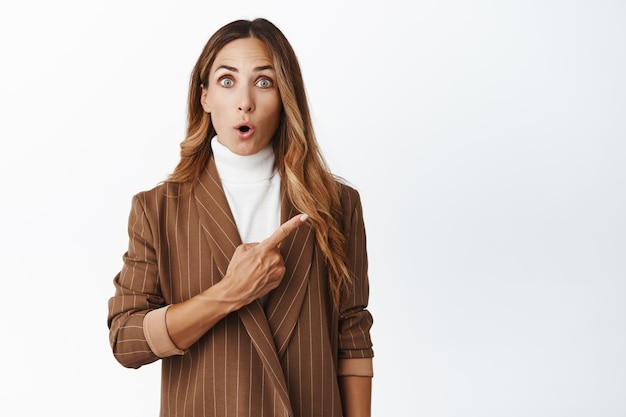 Portrait of surprised businesswoman gasping fascinated pointing at upper right corner promo showing discount or company brand name white background