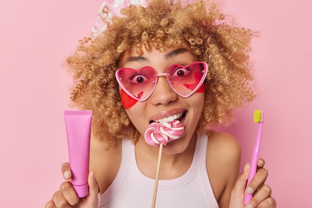 Portrait of surprised cheerful woman with curly bushy hair keeps lollipop in mouth holds toothpaste and toothbrush applies beauty patches under eyes wears pink heart sunglasses poses indoor