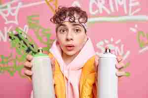 Foto gratuita ritratto di ragazzo bello hipster dai capelli ricci sorpreso utilizza spray aerosol per disegnare segni di graffiti luogo pubblico sembra meravigliato alla telecamera pone contro il muro dipinto colorato