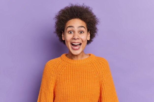 Portrait of surprised curly haired woman keeps mouth widely opened reacts on awesome surprise has overjoyed expression dressed in orange knitted sweater.