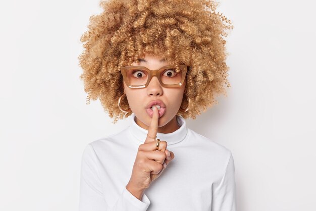 Portrait of surprised curly haired young woman makes silence gesture keeps index finger over lips demands complete silenece wears spectacles and casual jumper isolated over white background.